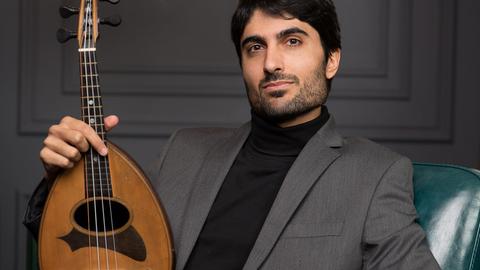 Ein Mann in grauem Anzug sitzt in einem grünen Ledersessel unter einem Kronleuchter und präsentiert seine Mandoline aufrecht auf dem Knie.