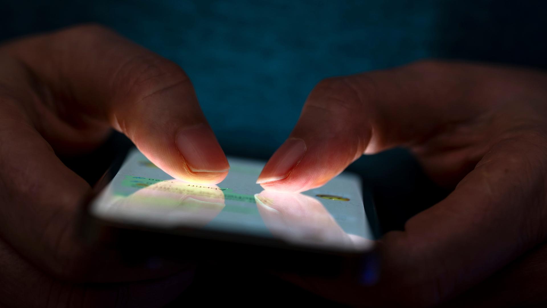 Eine Frau tippt auf ihrem Telefon. Die Beleuchtung hebt die Finger des Benutzers und den Bildschirm des Smartphones hervor.