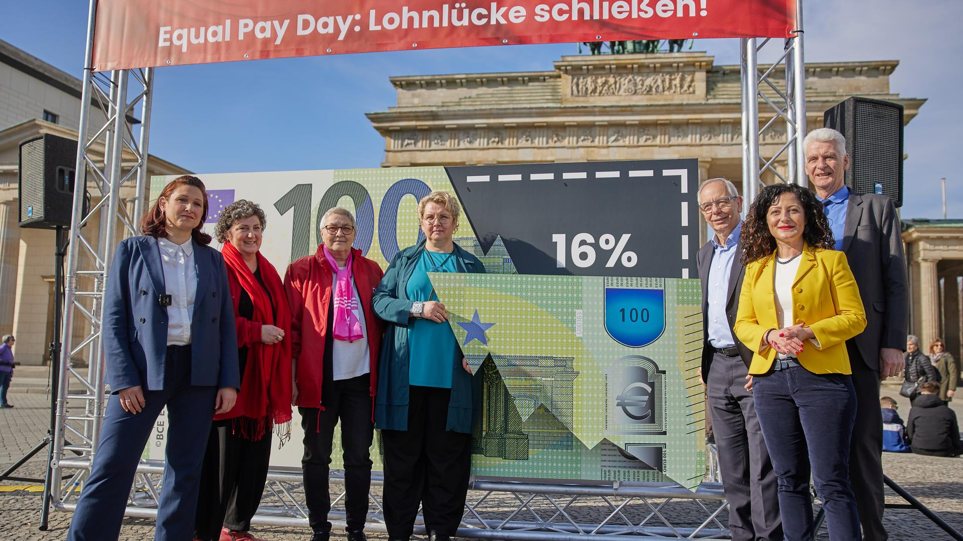 Anlässlich des Equal Pay Day steht Ferda Ataman (l.), Antidiskriminierungsbeauftragtedes Bundes, mit anderen am Brandenburger Tor in Berlin vor einem Plakat, das die Entgeltlücke von 16 Prozent zwischen Männern und Frauen darstellt.