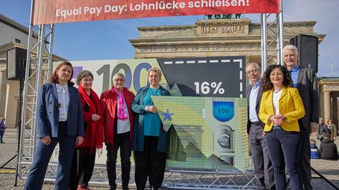 Anlässlich des Equal Pay Day steht Ferda Ataman (l.), Antidiskriminierungsbeauftragtedes Bundes, mit anderen am Brandenburger Tor in Berlin vor einem Plakat, das die Entgeltlücke von 16 Prozent zwischen Männern und Frauen darstellt.