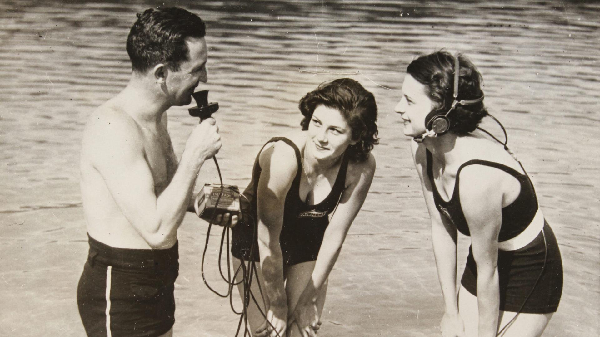 Auf dem Schwarzweißfoto von April 1934 ist links ein Mann in Badehose zu sehen, der ein Mikrofon hält und knietief im Wasser steht. Rechts von ihm stehen zwei junge Frauen, von denen eine einen Kopfhörer trägt, der über Kabel und ein Kästchen mit dem Mikro des Mannes verbunden ist. Die Frauen stehen auch im Wasser und schaeun den Mann an. 