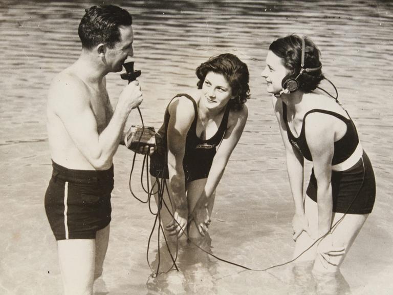 Auf dem Schwarzweißfoto von April 1934 ist links ein Mann in Badehose zu sehen, der ein Mikrofon hält und knietief im Wasser steht. Rechts von ihm stehen zwei junge Frauen, von denen eine einen Kopfhörer trägt, der über Kabel und ein Kästchen mit dem Mikro des Mannes verbunden ist. Die Frauen stehen auch im Wasser und schaeun den Mann an. 
