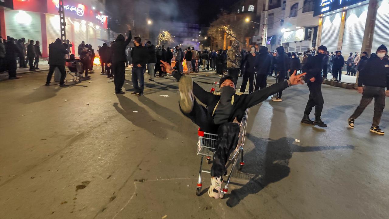 Das Bild zeigt viele Demonstranten im Dunkeln in einer Fußgängerzone. Im Vordergrund sitzt ein maskierter junger Mann in einem Einkaufswagen und steckt alle Arme und Beine von sich. Das Bild zeigt viele Demonstranten im Dunkeln in einer Fußgängerzone. Im Vordergrund sitzt ein maskierter junger Mann in einem Einkaufswagen und steckt alle Arme und Beine von sich.