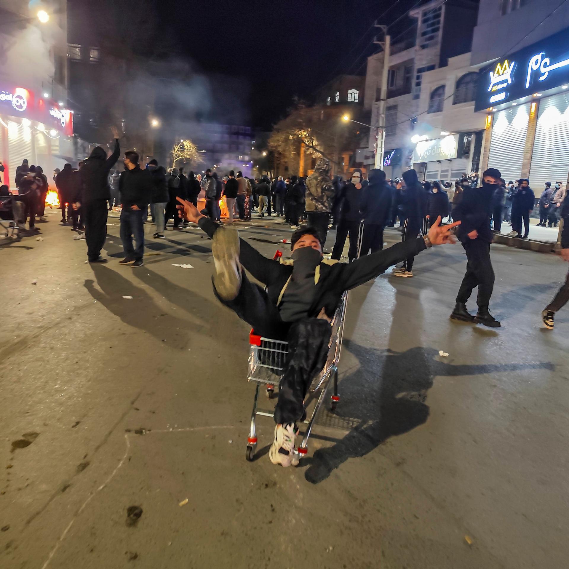 Das Bild zeigt viele Demonstranten im Dunkeln in einer Fußgängerzone. Im Vordergrund sitzt ein maskierter junger Mann in einem Einkaufswagen und steckt alle Arme und Beine von sich. 