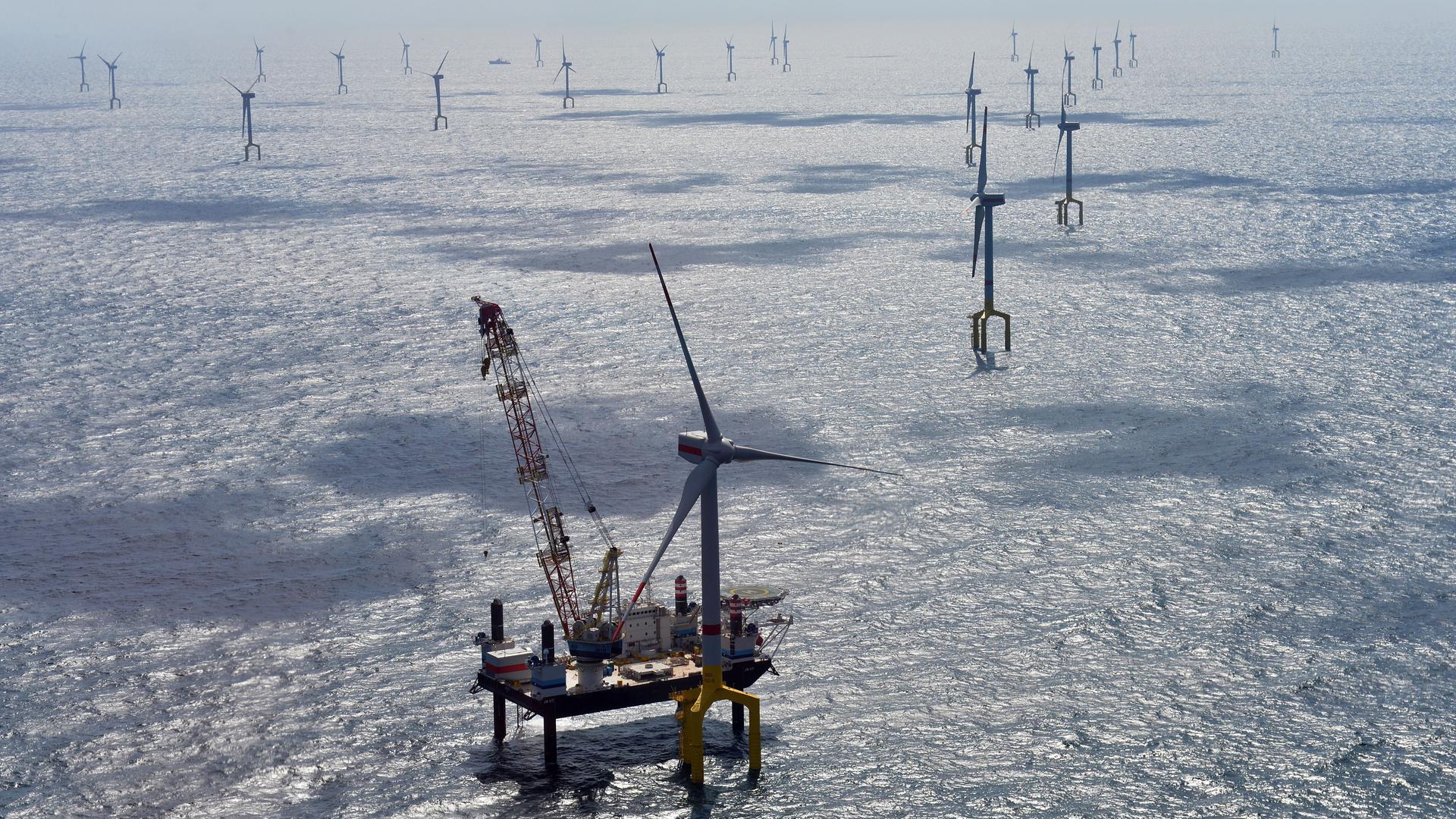 Luftaufnahme eines Hochseewindparks nordwestlich der Insel Borkum. Vor einem der Windräder schwimmt ein Spezialschiff im Wasser, von dem aus Arbeiten an dem Windrad verrichtet werden.