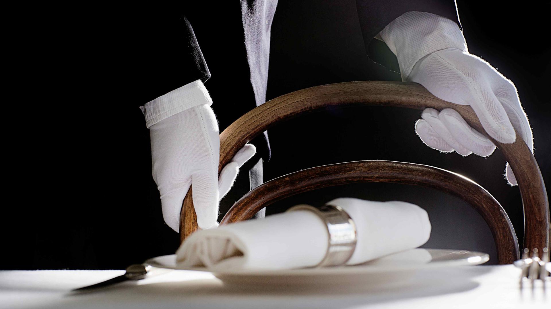Nahaufnahme eines edel gedecktes Tischplatzes, dahinter die Hände eines Kellners in weißen Handschuhen und schwarzem Sakko.