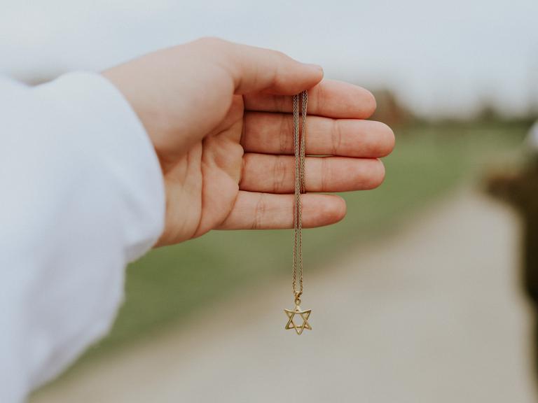 Nahaufnahme einer Hand, die eine goldene Halskette mit einem Anhänger in Form eines Davidsterns hält.