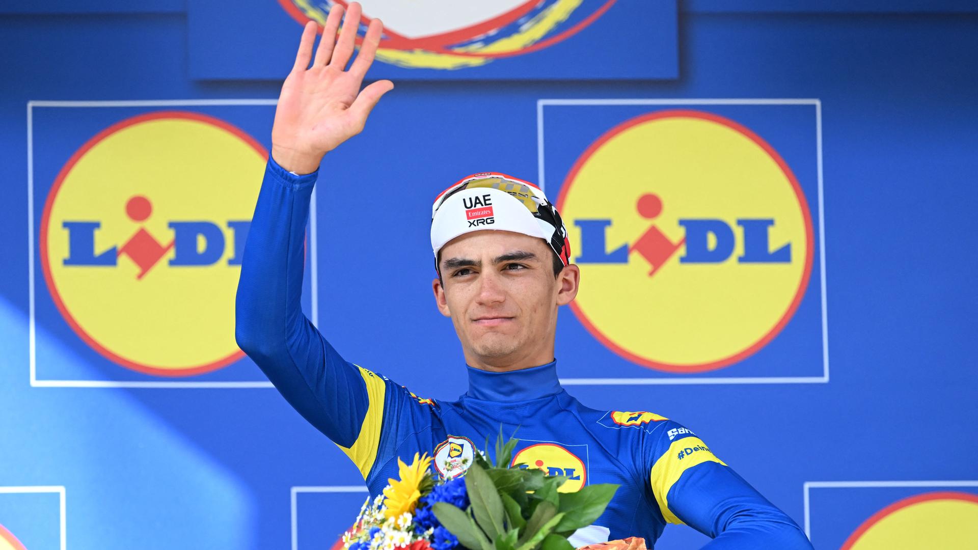 Radsport: Deutschland Tour - Der Mexikaner Isaac del Toro bejubelt seinen Gesamtsieg. Er hält einen Blumenstrauß in der einen Hand und winkt mit der anderen.