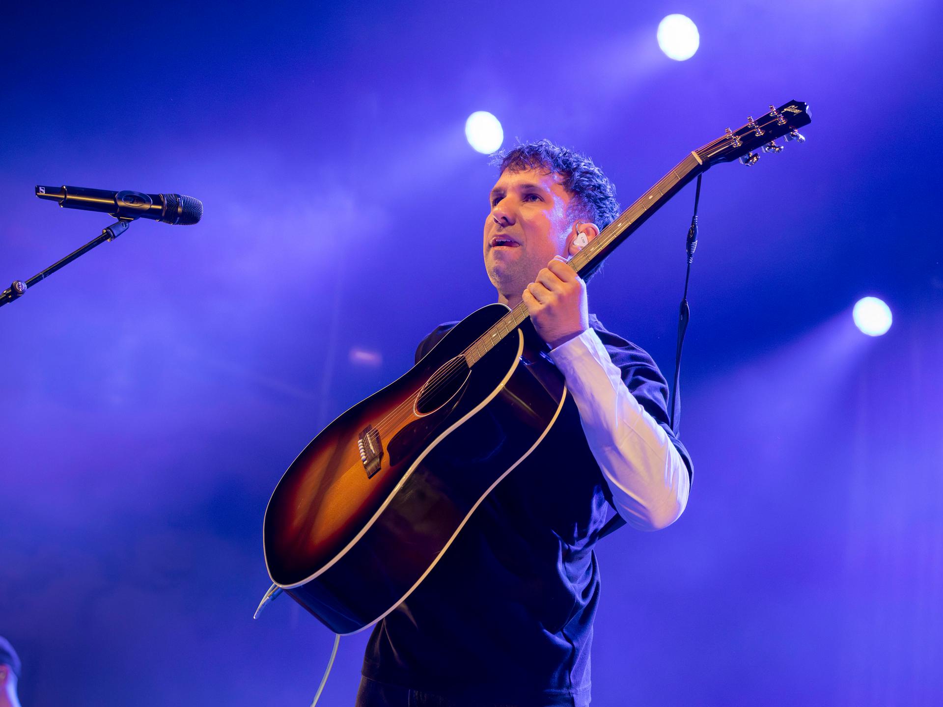 Der Musiker Betterov steht mit Gitarre auf einer Bühne vor einem Mikrofon und blickt ins Publikum. Der Hintergrund ist in violetten Farben ausgeleuchtet