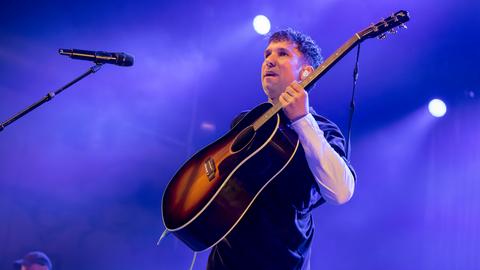 Der Musiker Betterov steht mit Gitarre auf einer Bühne vor einem Mikrofon und blickt ins Publikum. Der Hintergrund ist in violetten Farben ausgeleuchtet