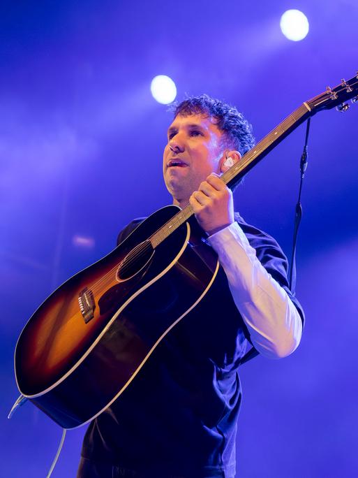 Der Musiker Betterov steht mit Gitarre auf einer Bühne vor einem Mikrofon und blickt ins Publikum. Der Hintergrund ist in violetten Farben ausgeleuchtet
