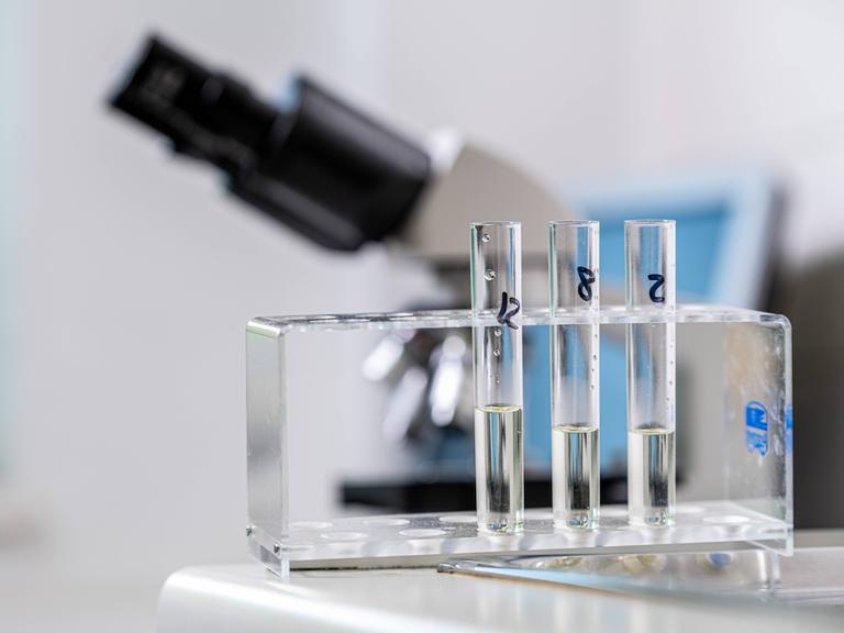 Drei Reagenzgläser im Labor mit einer klaren Flüssigkeit. Im Hintergrund ist ein Mikroskop erkennbar. Drei Reagenzgläser im Labor mit einer klaren Flüssigkeit. Im Hintergrund ist ein Mikroskop erkennbar.