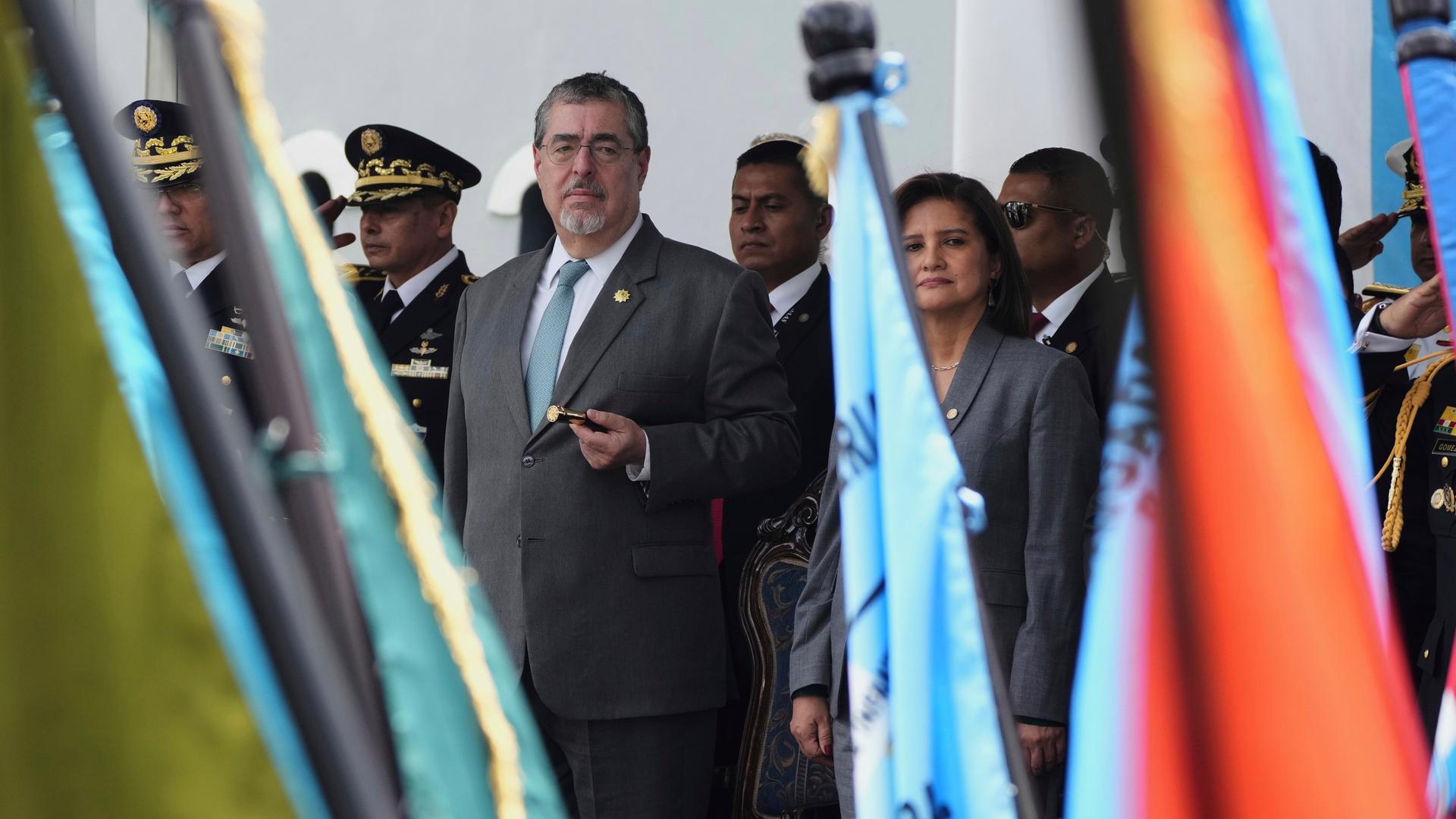 Bernardo Arévalo (Mitte) und Karin Herrera stehen neben hochrangigen Militärs auf einer Tribüne und blicken auf eine Militärparade mit vielen Fahnen