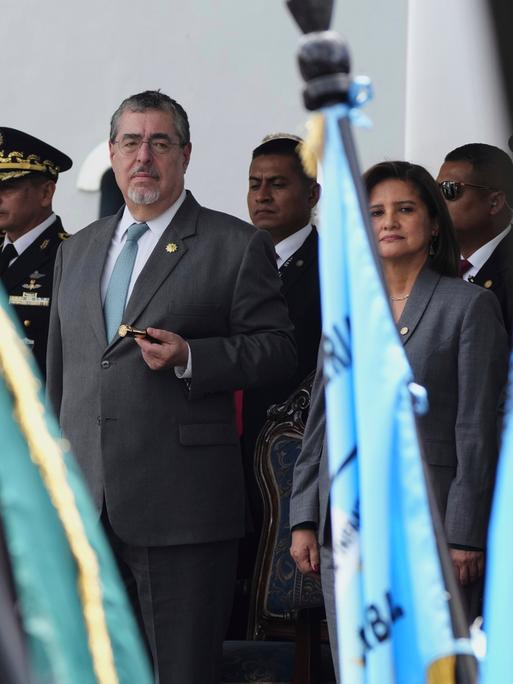 Bernardo Arévalo (Mitte) und Karin Herrera stehen neben hochrangigen Militärs auf einer Tribüne und blicken auf eine Militärparade mit vielen Fahnen