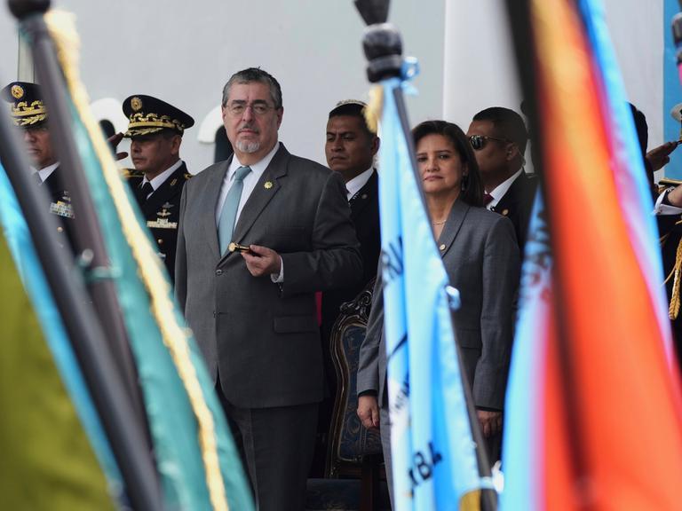 Bernardo Arévalo (Mitte) und Karin Herrera stehen neben hochrangigen Militärs auf einer Tribüne und blicken auf eine Militärparade mit vielen Fahnen