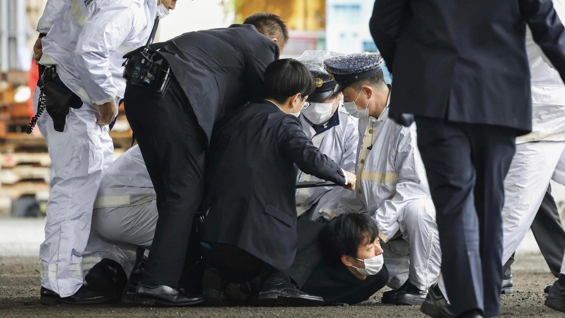Der Täter mit Schutzmaske liegt am Boden und schaut nach oben. Um ihn herum Polizisten in weißen Uniformen und Sicherheitsleute in schwarzen Anzügen, die ihn festhalten.
