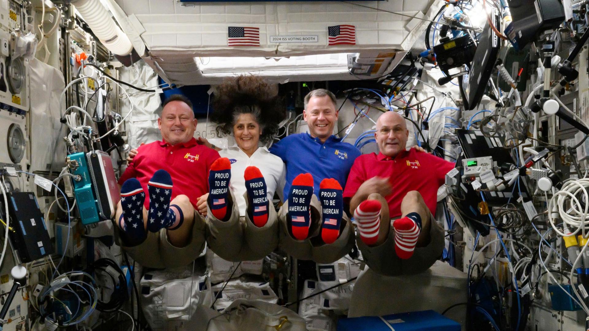 Die Astronauten Butch Wilmore, Suni Williams, Nick Hague and Don Pettit schweben und strecken ihre Füße mit US-Socken nach vorne. 