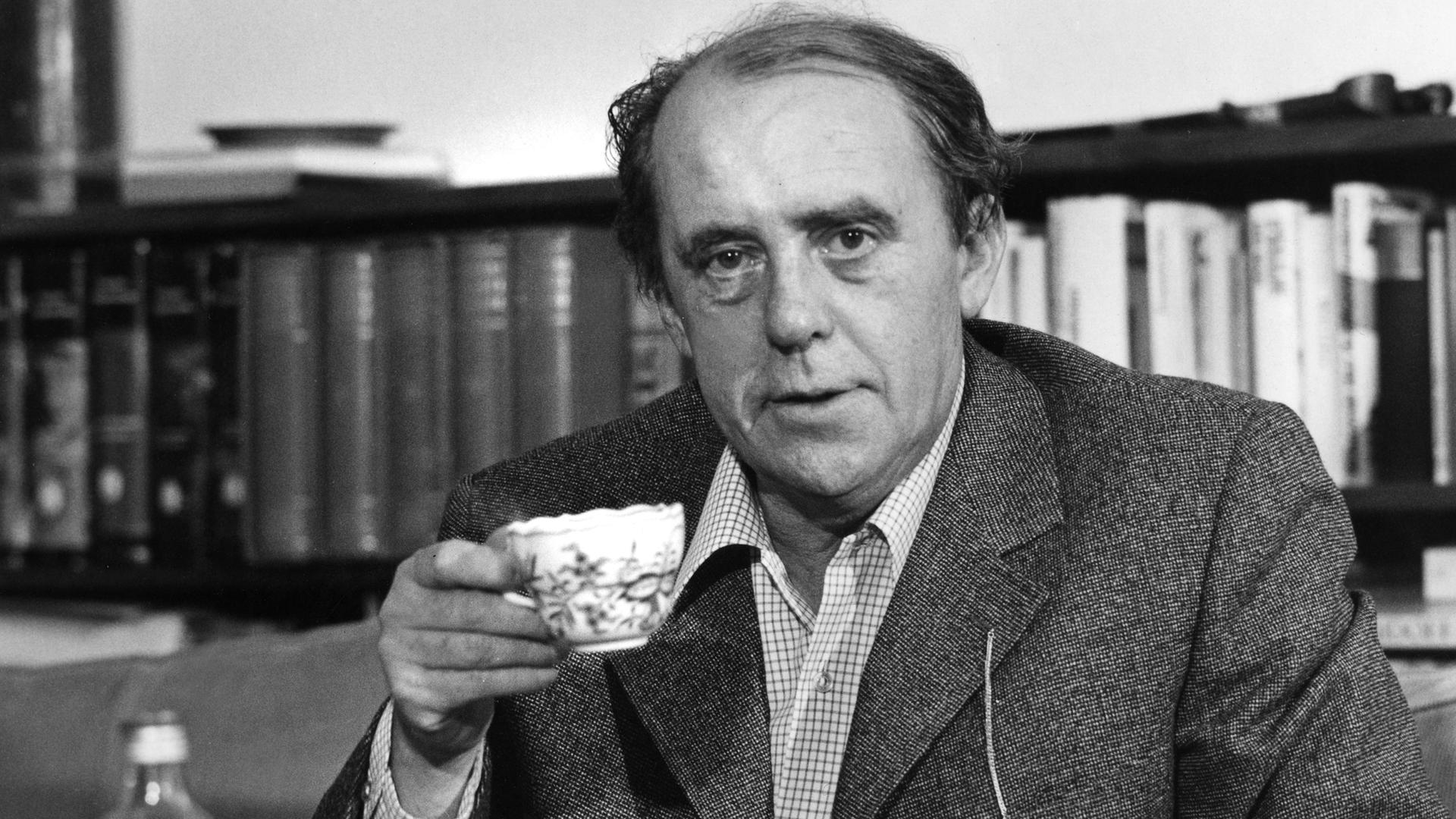 Schwarzweißaufnahme von Heinrich Böll, der mit Kaffeetasse und Zigarette in seiner Wohnung an einem Kaffeetisch sitzt.