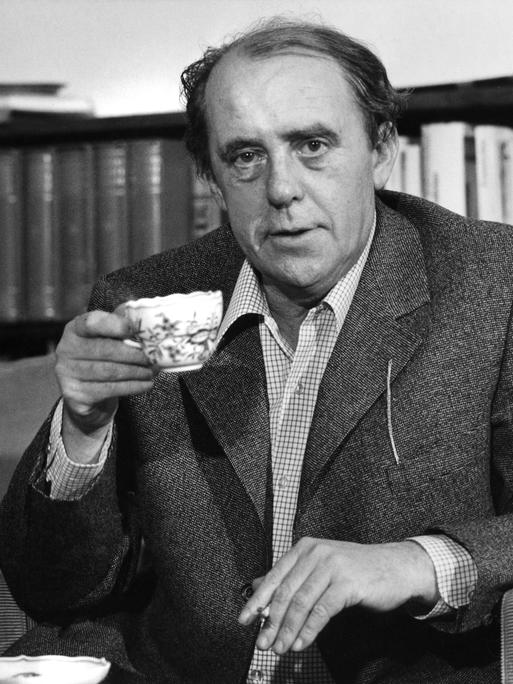 Schwarzweißaufnahme von Heinrich Böll, der mit Kaffeetasse und Zigarette in seiner Wohnung an einem Kaffeetisch sitzt.