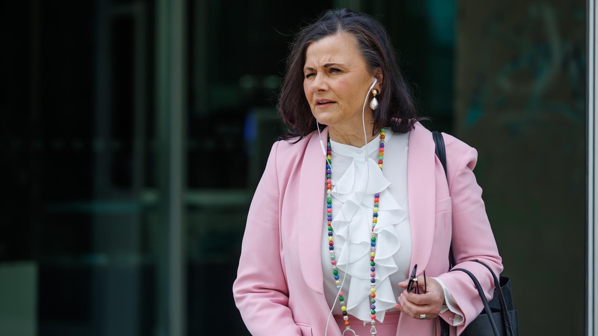 Connemann steht im Freien vor einer Glasfront. Sie trägtb ein rosa Jackett und hat Kopfhörer in den Ohren.