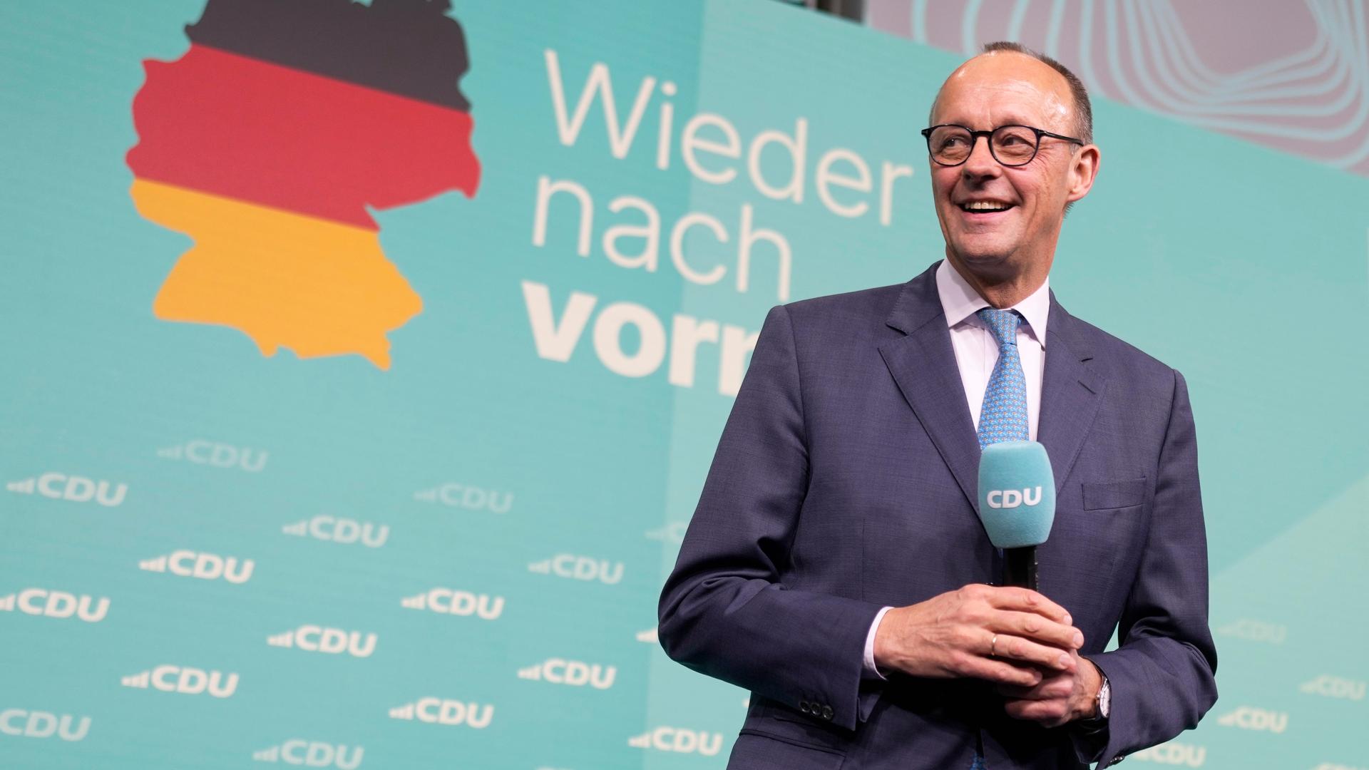 CDU-Chef Friedrich Merz freut sich am Wahlabend. Hinter ihm eine Deutschlandkarte und der Slogan "Wieder nach vorn".  Er hält ein Mikro mit CDU in der Hand. 