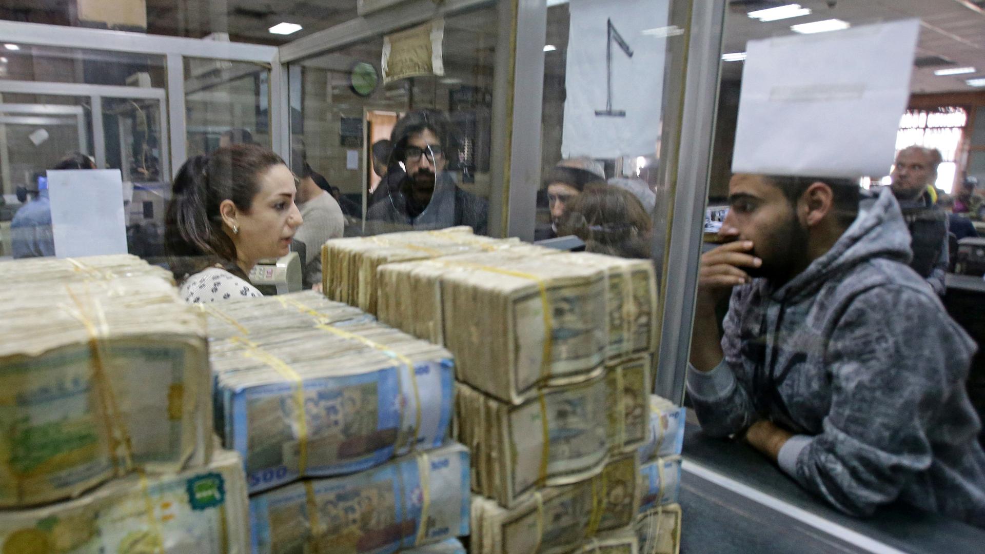 Syrer warten darauf, bei einer Bank in Damaskus Geld abheben zu können. Am Schalter liegen Bündel von Geldscheinen. 