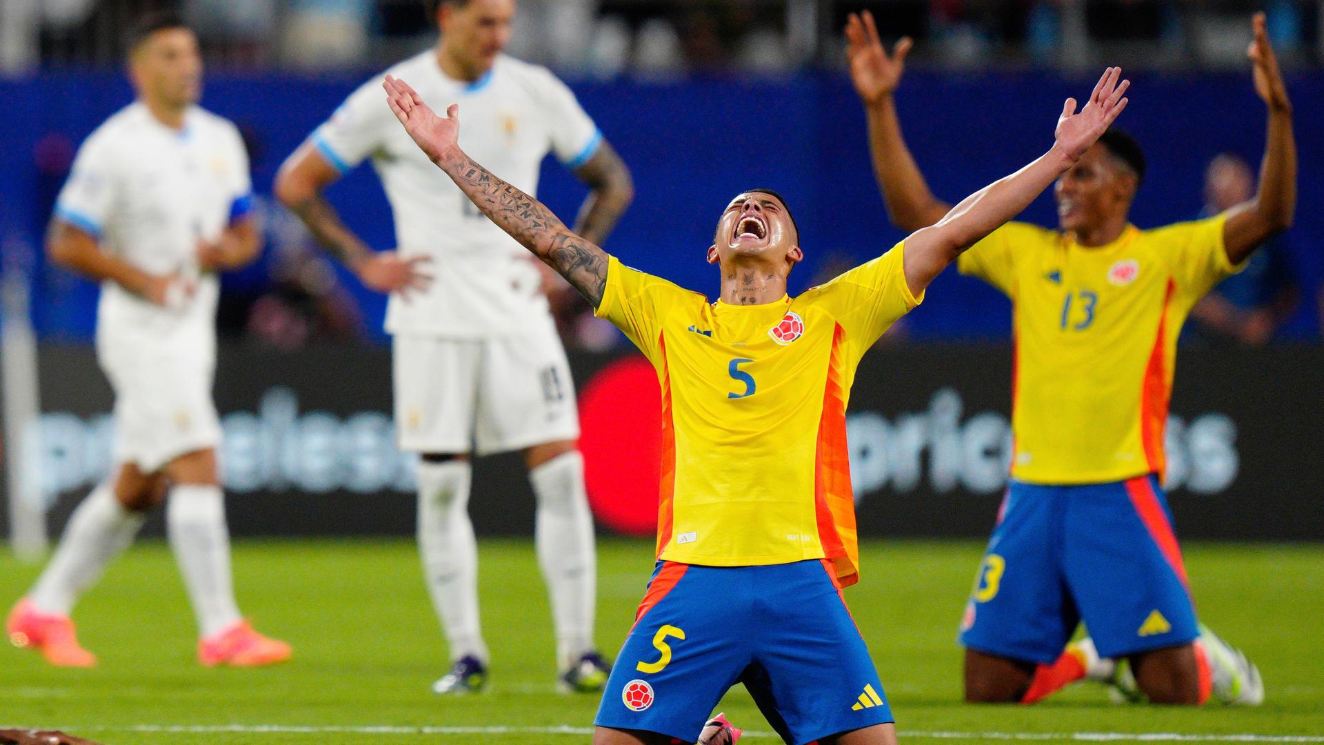 Zwei kolumbianische Spieler knien auf dem Rasen und halten die Hände jubelnd in die Höhe. Hinter ihnen stehen verschwommen zwei enttäuschte Spieler von Uruguay.