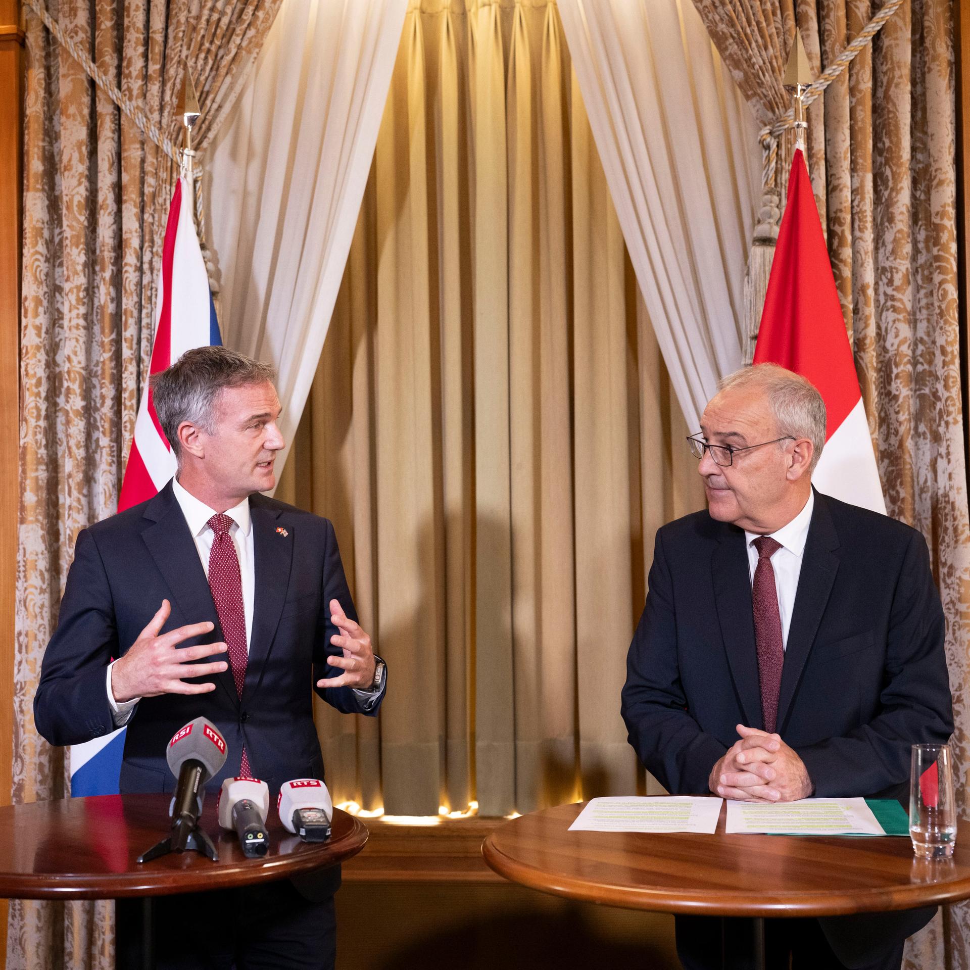 Der britische Handelsminister Kyle und der Schweizerische Bundespräsident Parlemin stehen jeweils an Rednerpulten nebeneinander.