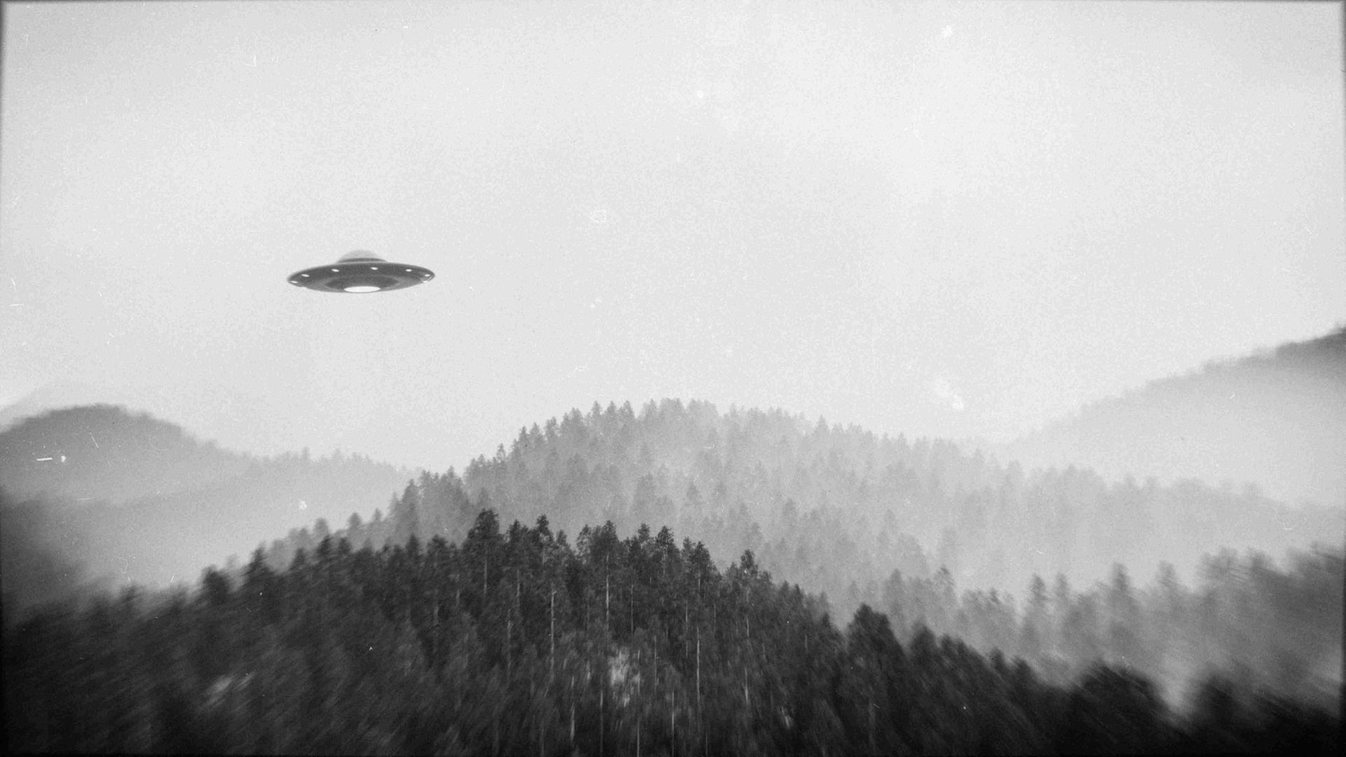 Schwarz-Weiß-Bild: Ein fliegendes Ufo über einer Waldlandschaft.