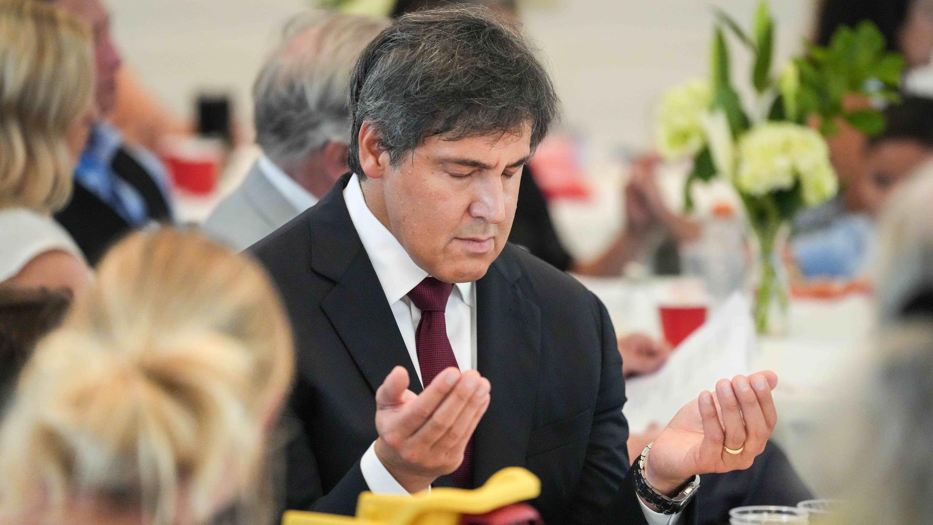 Ein Mann in Anzug hebt während eines Gebets die geöffneten Hände nach oben. Er senkt den Kopf und hat die Augen geschlossen. 