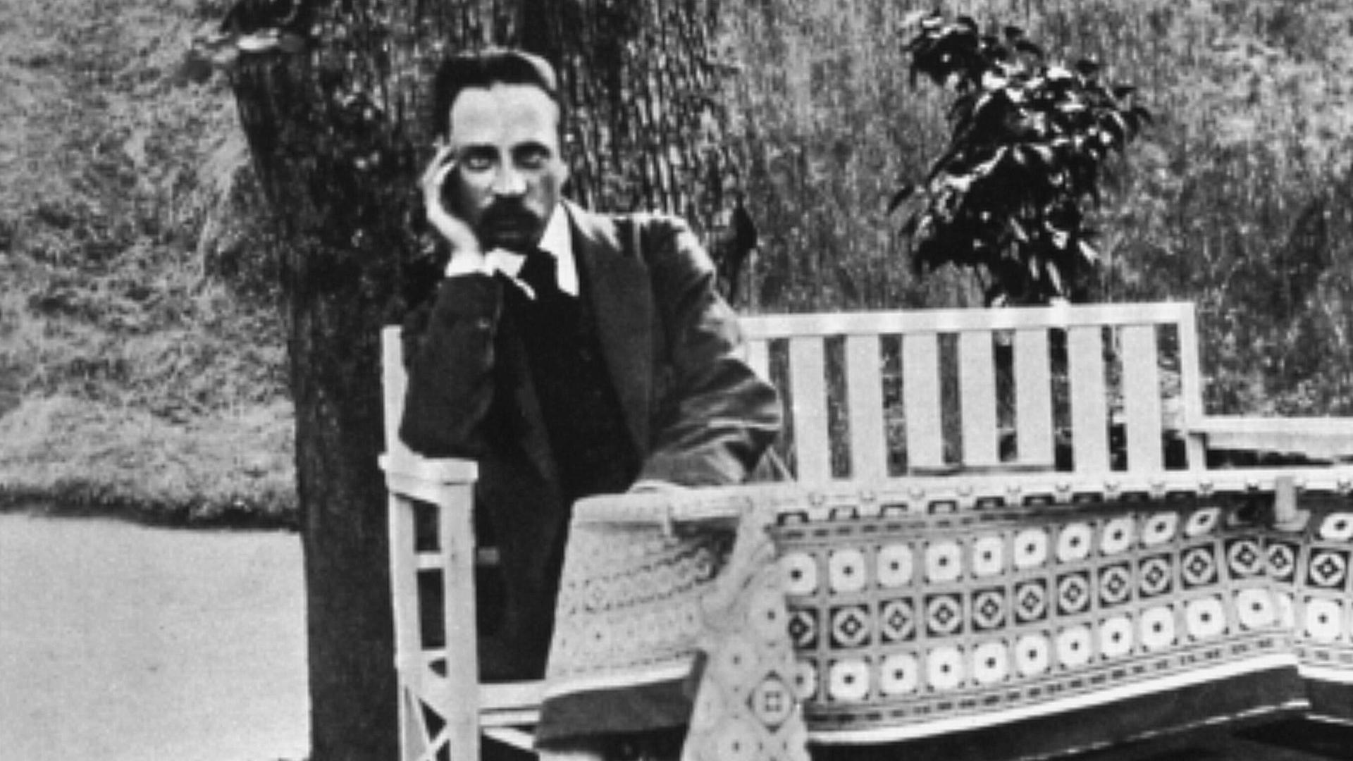 Der Dichter Rainer Maria Rilke sitzt auf einer weißen Gartenbank vor einem Baum im Jahr 1913.