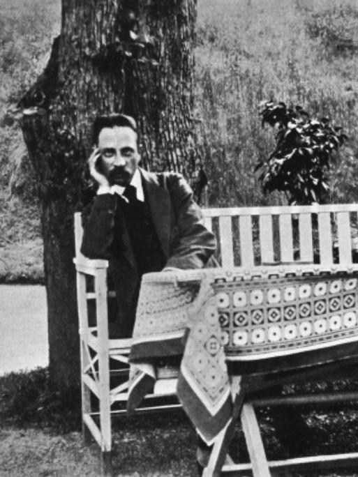 Der Dichter Rainer Maria Rilke sitzt auf einer weißen Gartenbank vor einem Baum im Jahr 1913.