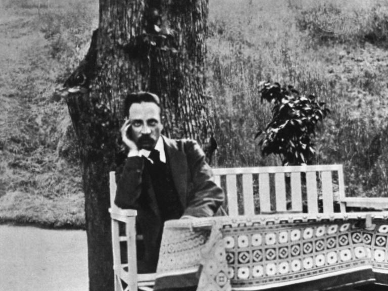 Der Dichter Rainer Maria Rilke sitzt auf einer weißen Gartenbank vor einem Baum im Jahr 1913.