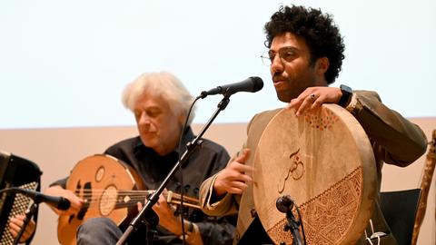 Mohcine Ramdan bei einem Auftritt mit seiner Band. Er singt in ein Mikrofon und spielt eine große flache Trommel. Mohcine Ramdan bei einem Auftritt mit seiner Band. Er singt in ein Mikrofon und spielt eine große flache Trommel.