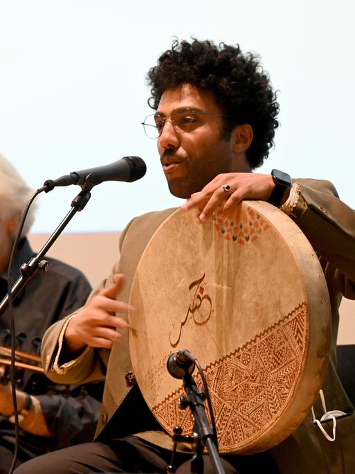 Mohcine Ramdan bei einem Auftritt mit seiner Band. Er singt in ein Mikrofon und spielt eine große flache Trommel.