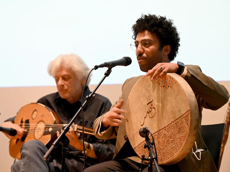 Mohcine Ramdan bei einem Auftritt mit seiner Band. Er singt in ein Mikrofon und spielt eine große flache Trommel.