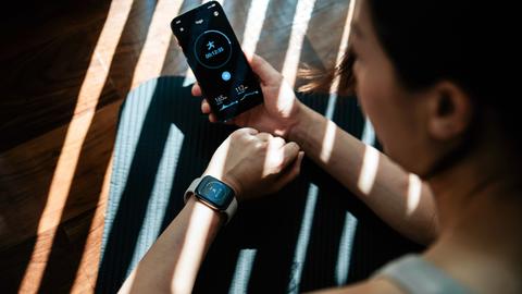 Eine junge Frau, die zu Hause trainiert und dabei Gesundheits- und Fitness-Apps auf ihrem Smartphone und ihrer Smartwatch am Handgelenk nutzt.