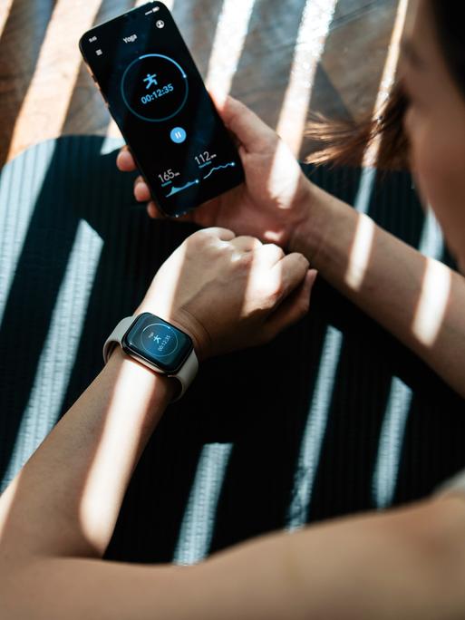 Eine junge Frau, die zu Hause trainiert und dabei Gesundheits- und Fitness-Apps auf ihrem Smartphone und ihrer Smartwatch am Handgelenk nutzt.
