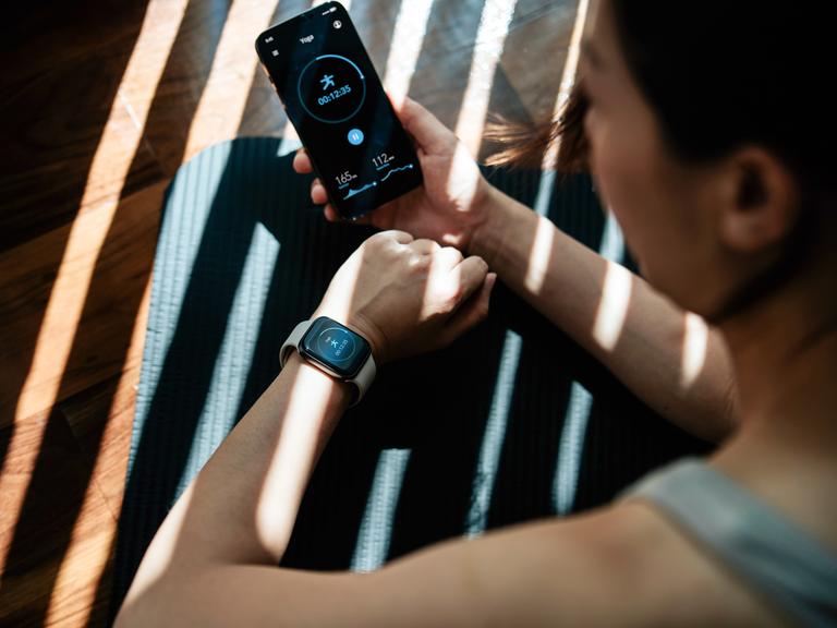 Eine junge Frau, die zu Hause trainiert und dabei Gesundheits- und Fitness-Apps auf ihrem Smartphone und ihrer Smartwatch am Handgelenk nutzt.