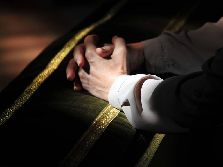 Auf einer Kirchenbank liegen die zum Gebet gefalteten Hände eines jungen Mannes.
