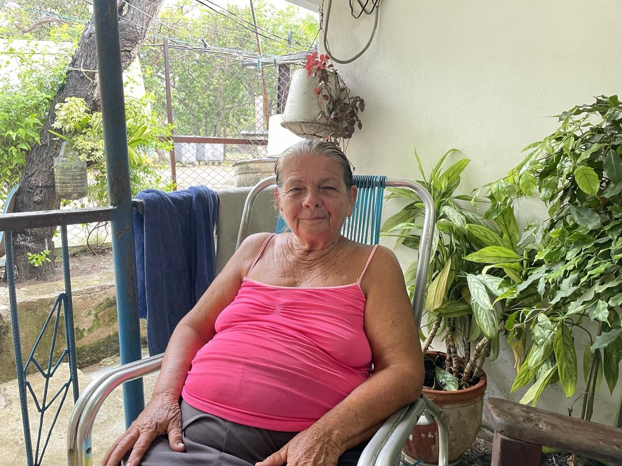 Martha Roldan, eine ältere Frau, sitzt in pinkem Trägershirt auf einem Stuhl auf einem Balkon