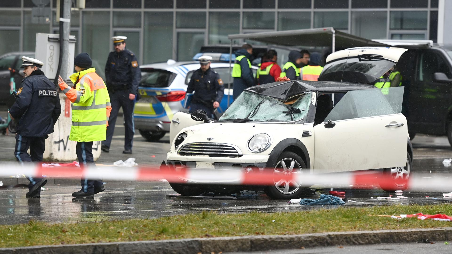 Ein beschädigtes beigefarbenes Auto steht in München auf der Straße, daneben sind Polizeibeamte zu sehen.