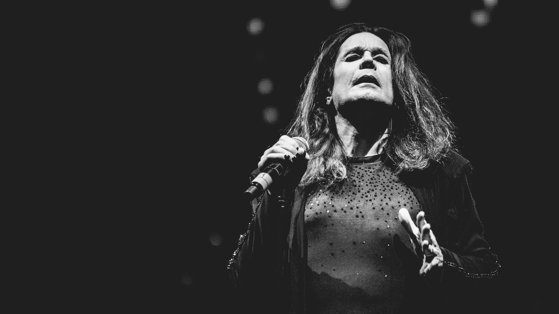 Schwarzweißaufnahme von Ozzy Osbourne, der in schwarzer Kleidung auf einer Bühne steht und so wirkt, als würde er einen Moment innehalten Schwarzweißaufnahme von Ozzy Osbourne, der in schwarzer Kleidung auf einer Bühne steht und so wirkt, als würde er einen Moment innehalten