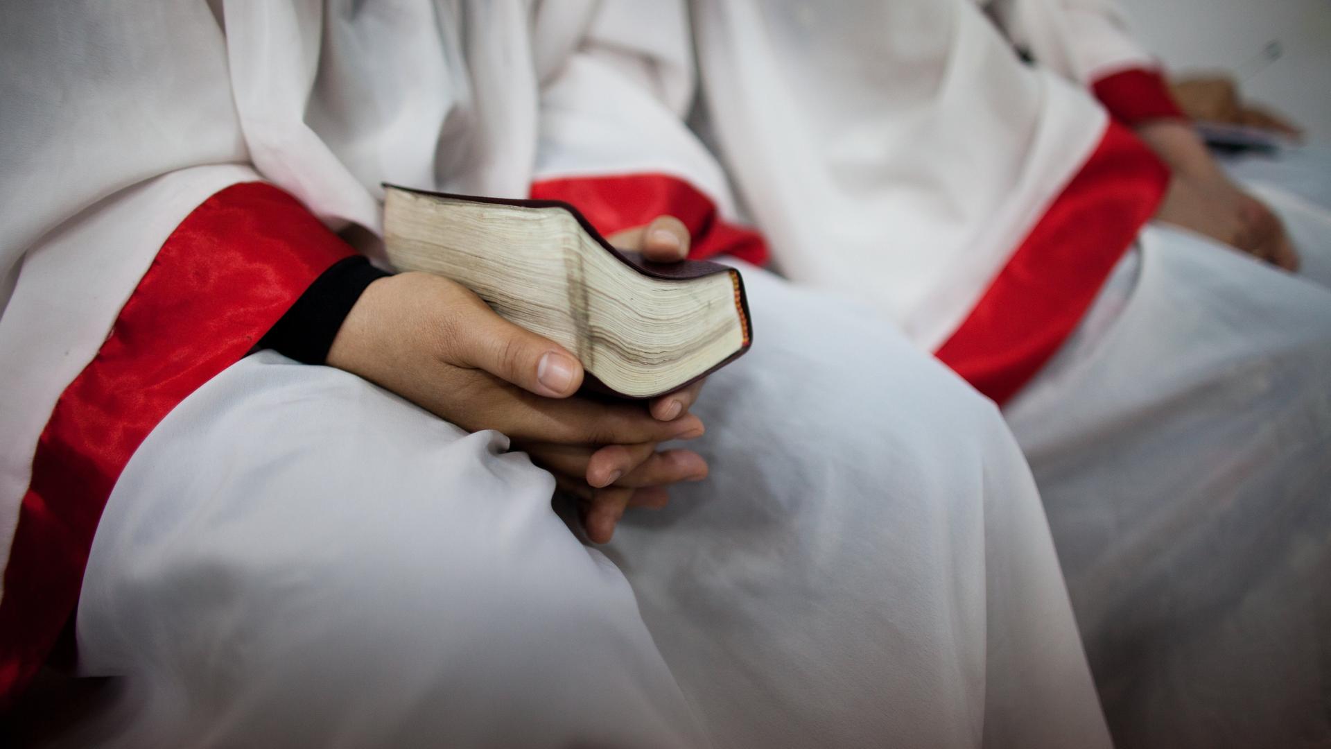 Eine Person in einem weißen Umhang mit roten Abnähern an den Armen hält eine Bibel im Schoß.