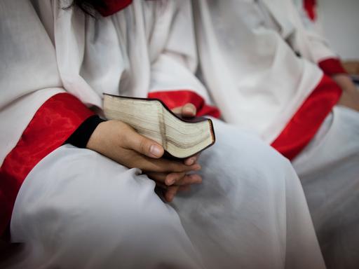 Eine Person in einem weißen Umhang mit roten Abnähern an den Armen hält eine Bibel im Schoß. Eine Person in einem weißen Umhang mit roten Abnähern an den Armen hält eine Bibel im Schoß.
