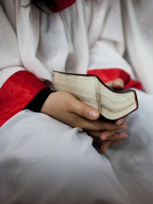 Eine Person in einem weißen Umhang mit roten Abnähern an den Armen hält eine Bibel im Schoß.