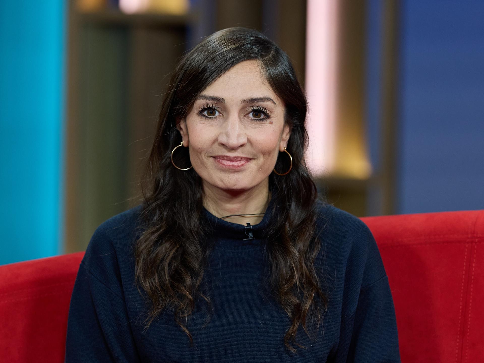 Die Journalistin und Autorin Gilda Sahebi sitzt  vor der Ausstrahlung von DAS! Rote Sofa, einer Live-Talksendung des Norddeutschen Rundfunks (NDR), auf einem roten Sofa und lächelt in die Kamera.