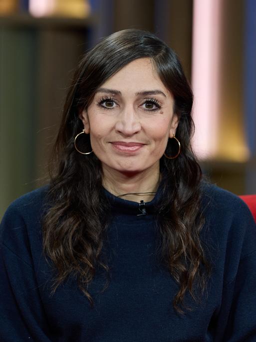 Die Journalistin und Autorin Gilda Sahebi sitzt  vor der Ausstrahlung von DAS! Rote Sofa, einer Live-Talksendung des Norddeutschen Rundfunks (NDR), auf einem roten Sofa und lächelt in die Kamera.