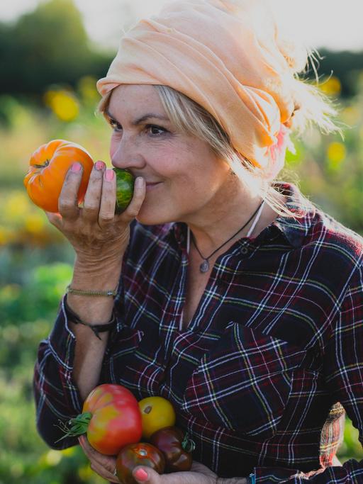 Eine Gärtnerin steht lächelnd auf einem Feld und riecht an einer frisch geernteten Tomate