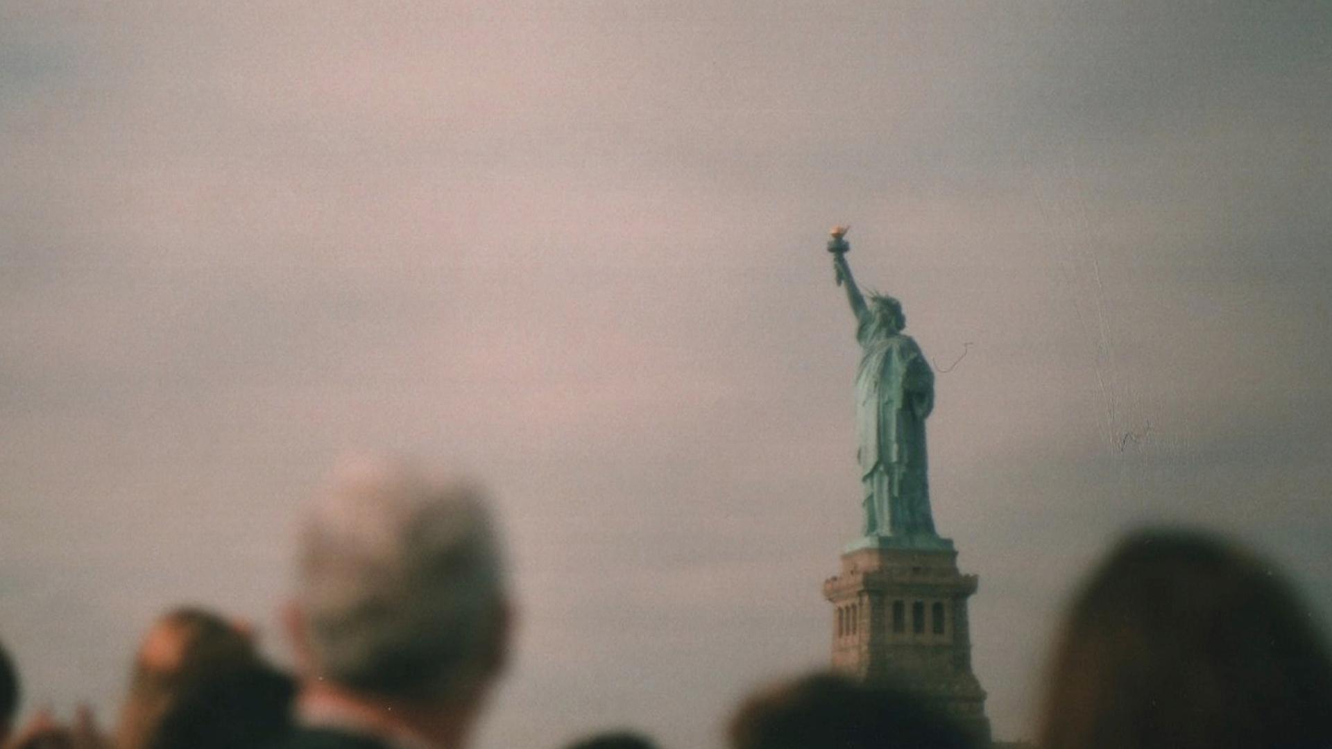 Menschen schauen auf die Freiheitsstatue, vor gräulich-flimmerndem Himmel.